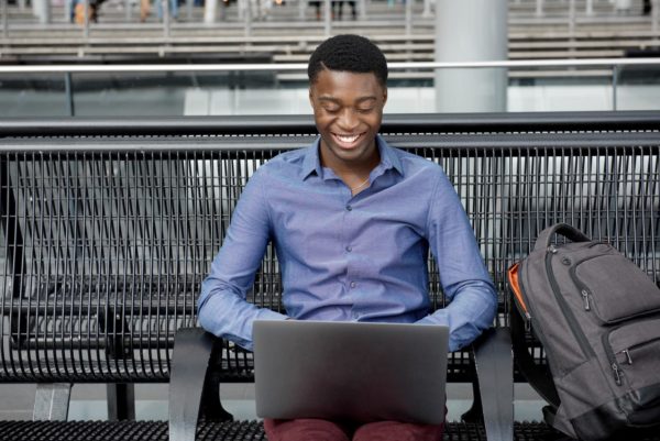 joven afroamericano sentado en un banco con bolsos y ordenador portátil